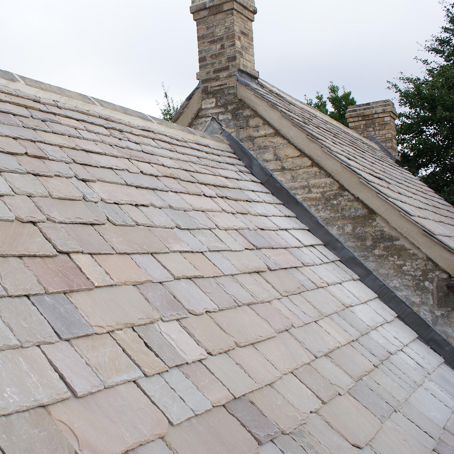 Cotswold Style Natural Stone Roof Slates (18mm) | Truestone | Truestone Slate roof with a stone chimney and a slate valley seam under an overcast sky.