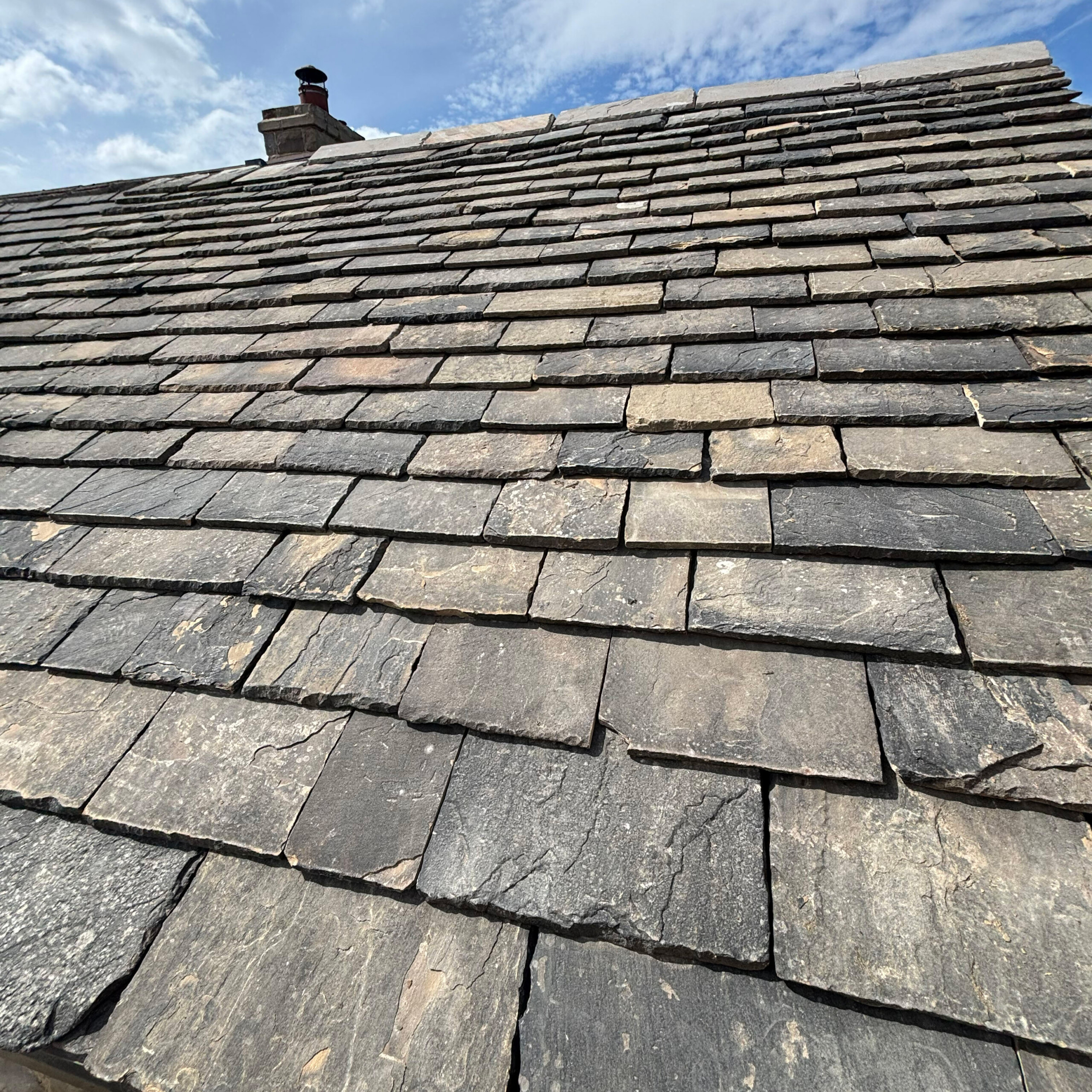 Reclaimed Yorkshire Stone Roof Slates | Truestone | Truestone Slate roof with layered gray shingles and a small chimney against a blue sky.