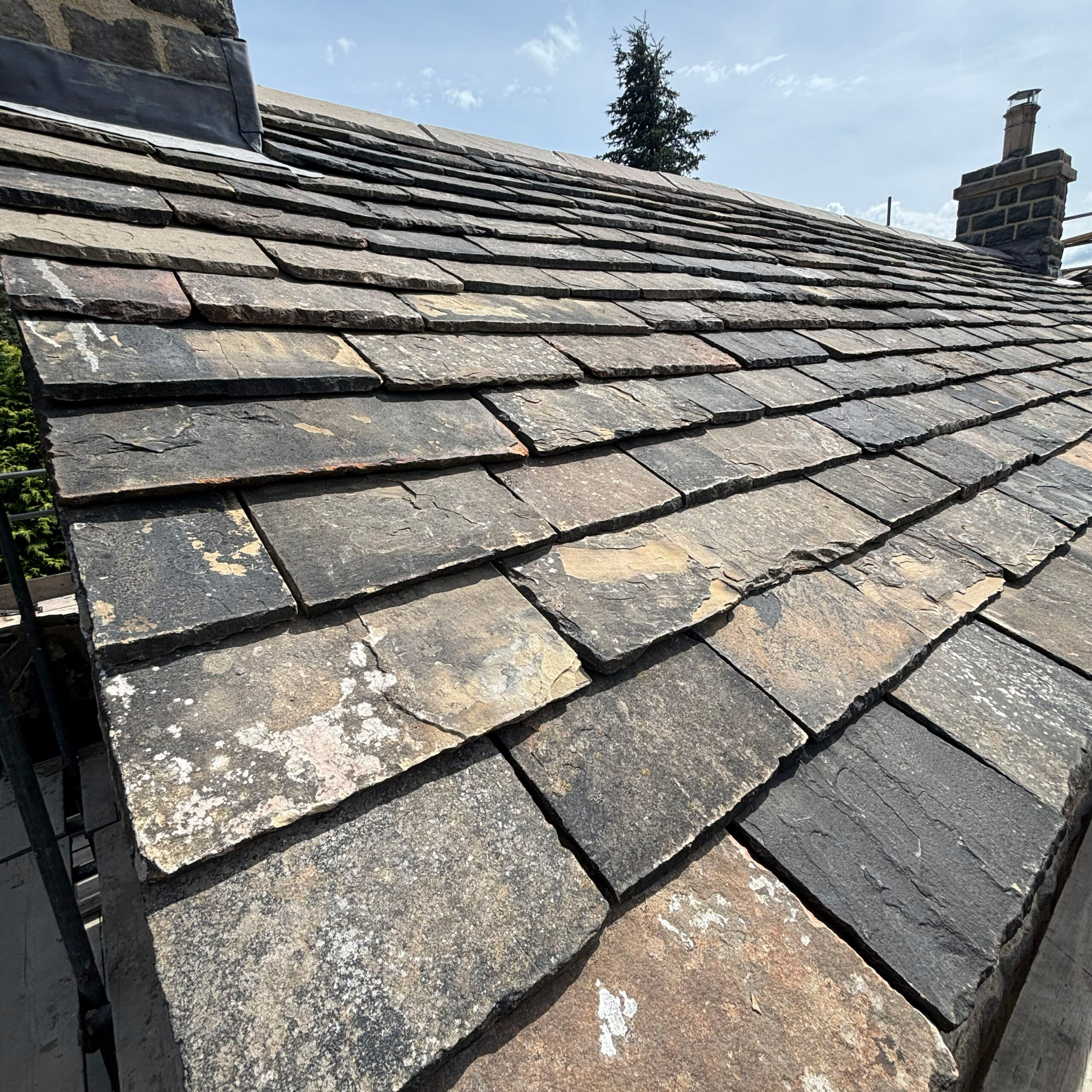 Reclaimed Yorkshire Stone Roof Slates | Truestone | Truestone Close-up of weathered slate roof tiles with a brick chimney visible on the right and a blue sky above.