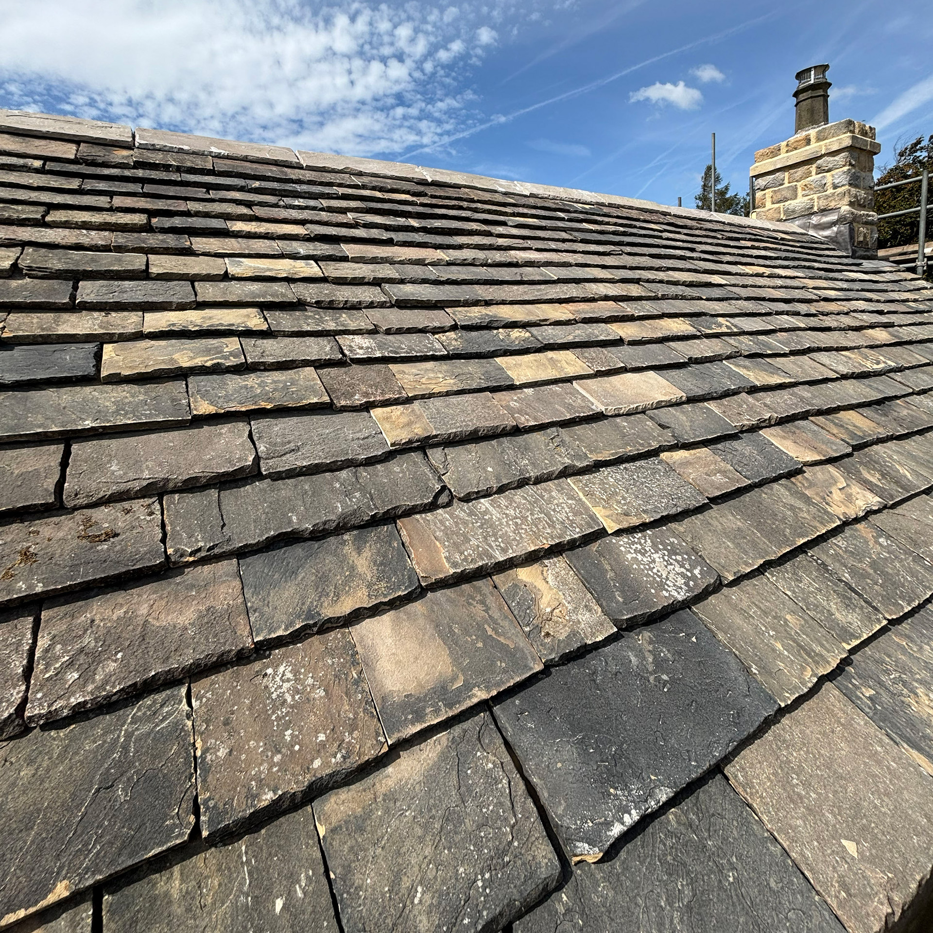 Reclaimed Yorkshire Stone Roof Slates | Truestone | Truestone Slate roof with overlapping dark gray shingles and a stone chimney against a blue sky.